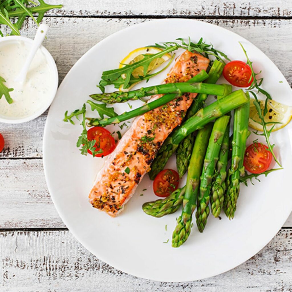 Lachs mit grünem Spargel und Kirschtomaten wird von oben auf einem weißen Teller präsentiert.