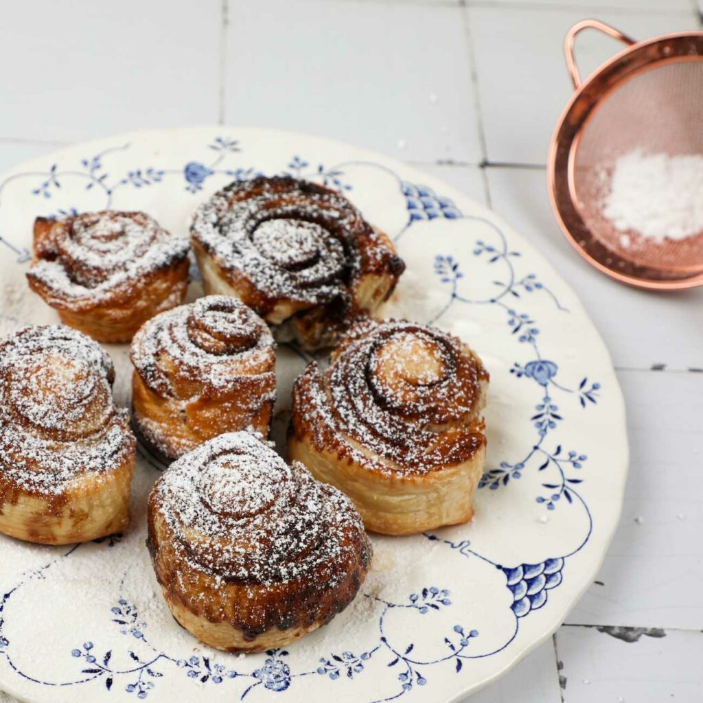 Zimtschnecken mit Blätterteig werden mit Puderzucker bestreut auf einem weiß-blauen Teller gezeigt.